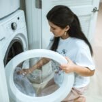 A woman kneeling to load clothes into a front-loading washing machine in a modern home laundry room.